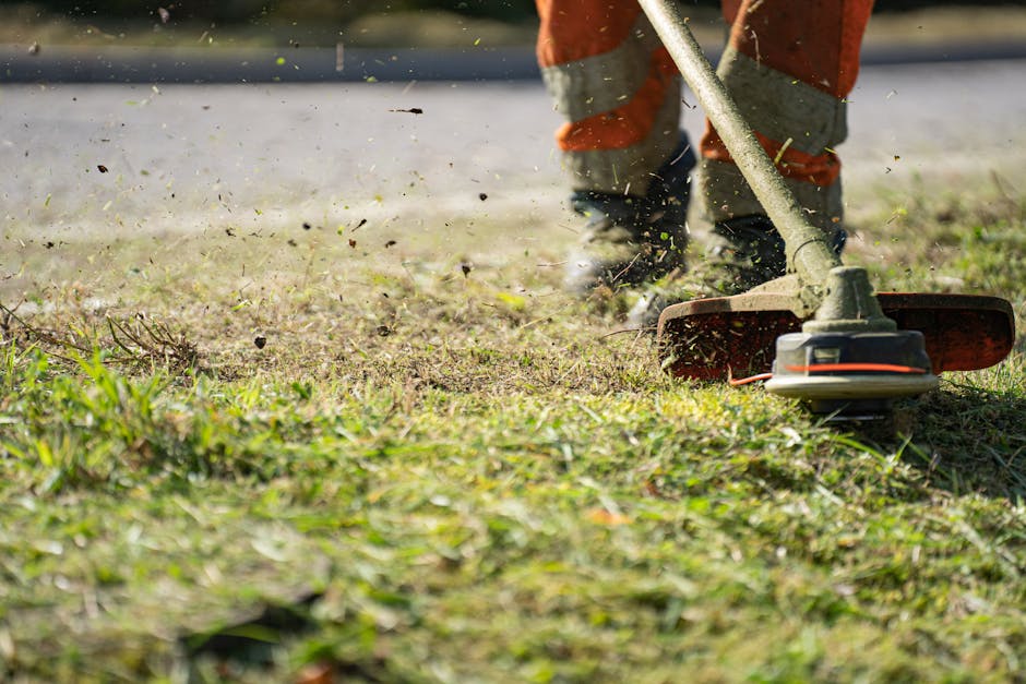 A gardener wearing orange work trousers is using a grass trimmer with a long handle and a rotary cutting head to maintain a lawn in a garden. The trimmer is actively cutting grass and small weeds, wit