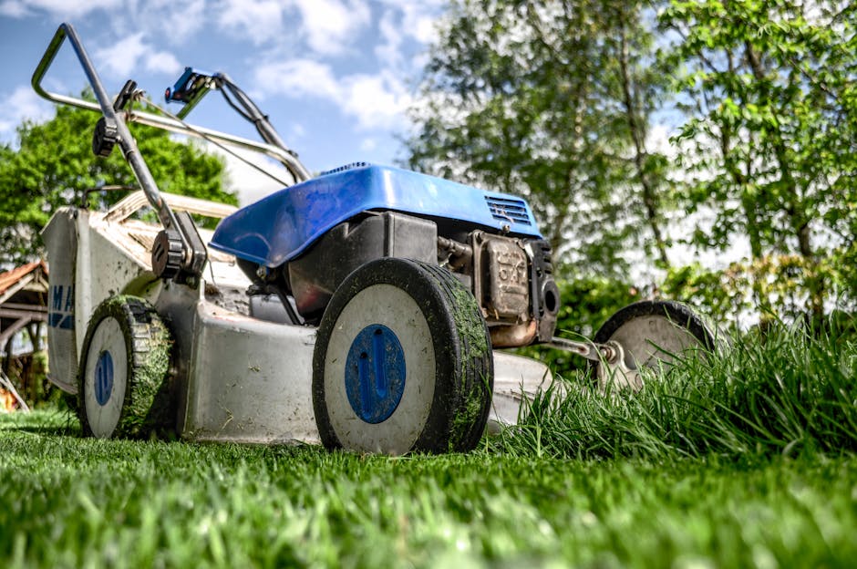 A gardener wearing orange work trousers is using a grass trimmer with a long handle and a rotary cutting head to maintain a lawn in a garden. The trimmer is actively cutting grass and small weeds, wit