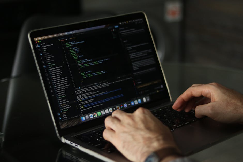 A person working on a sleek, modern laptop with a black keyboard, displaying lines of code in various colors on a dark background. The person's hands are positioned over the keyboard, with one hand's